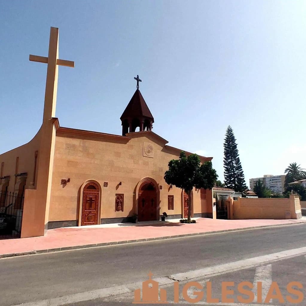 Exterior de la Iglesia Armenia Santa María Madre de Dios en Málaga: fachada de piedra clara, puertas de madera tallada con cruces, hornacinas decorativas, cúpula cónica y acera roja bajo cielo azul.