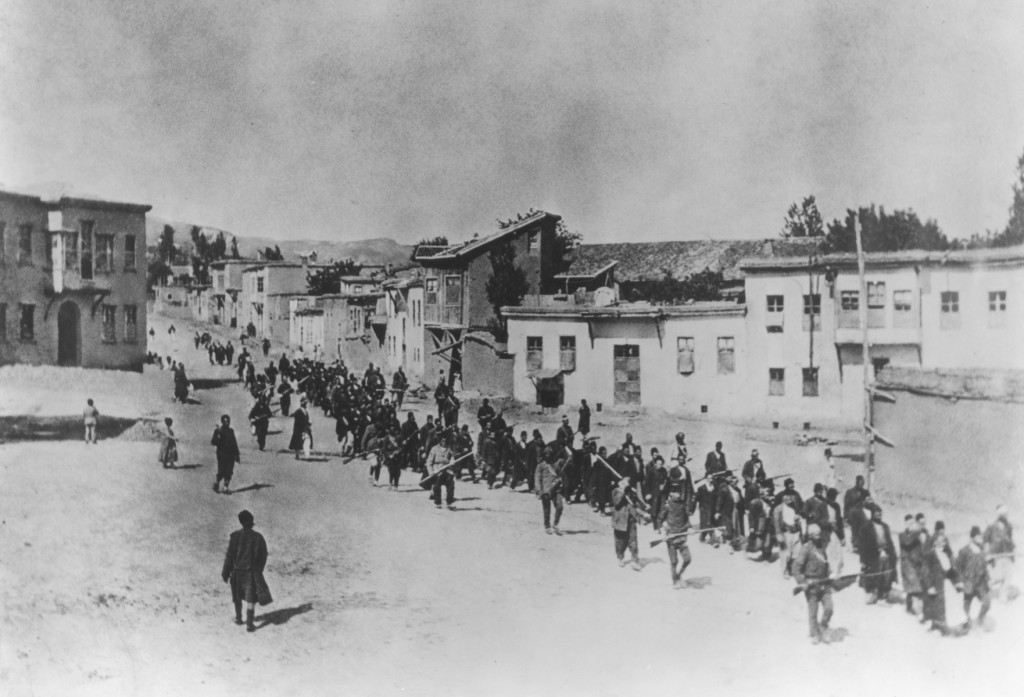 Fotografía en blanco y negro: larga columna de hombres civiles en calle de pueblo, escoltados por soldados con fusiles, 1915.