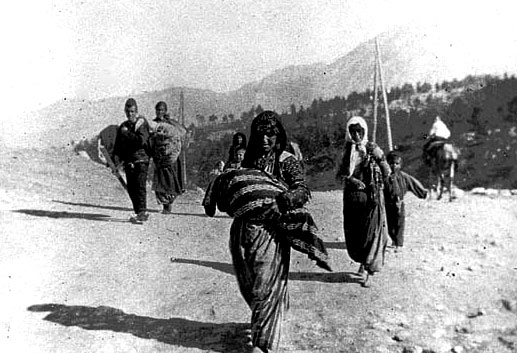 Grupo de refugiados, sobre todo mujeres y niños, caminando con hatos en paisaje árido y montañoso.