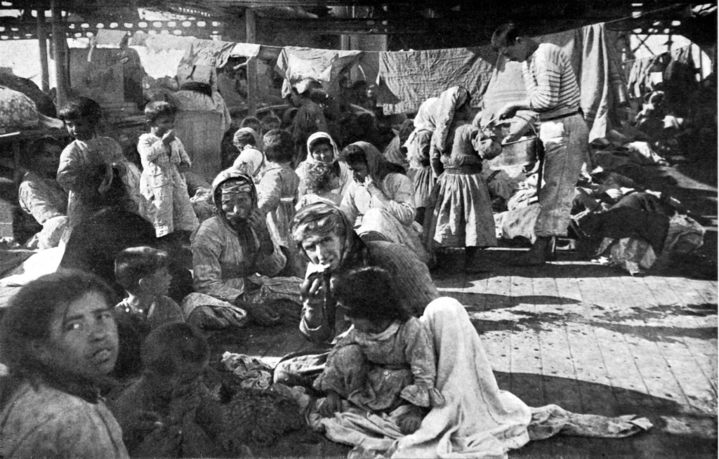 Cubiertas de buque con numerosas personas, ropa colgada y un marinista atiende a refugiados, 1915.