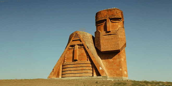 Monumento Nosotros somos nuestras montañas: cabezas monumentales de toba de anciano y anciana con pañuelo tradicional, rostros estilizados bajo cielo azul.