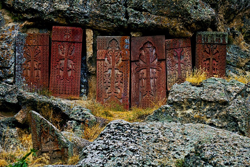 Fila de seis khachkares armenios—estelas de piedra con cruces centrales y relieves vegetales—bajo un voladizo rocoso natural; toba rojiza, peñasco gris y hierba seca.