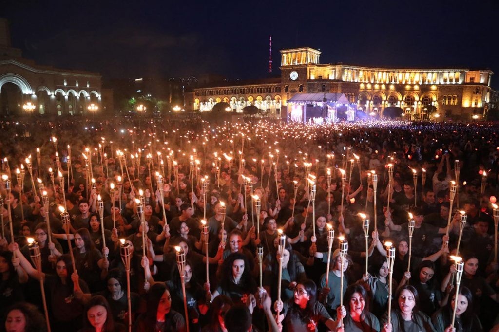 Miles de personas en una marcha nocturna con antorchas en la plaza de la República de Ereván en memoria del genocidio armenio, con la torre del reloj de la Casa de Gobierno iluminada al fondo.